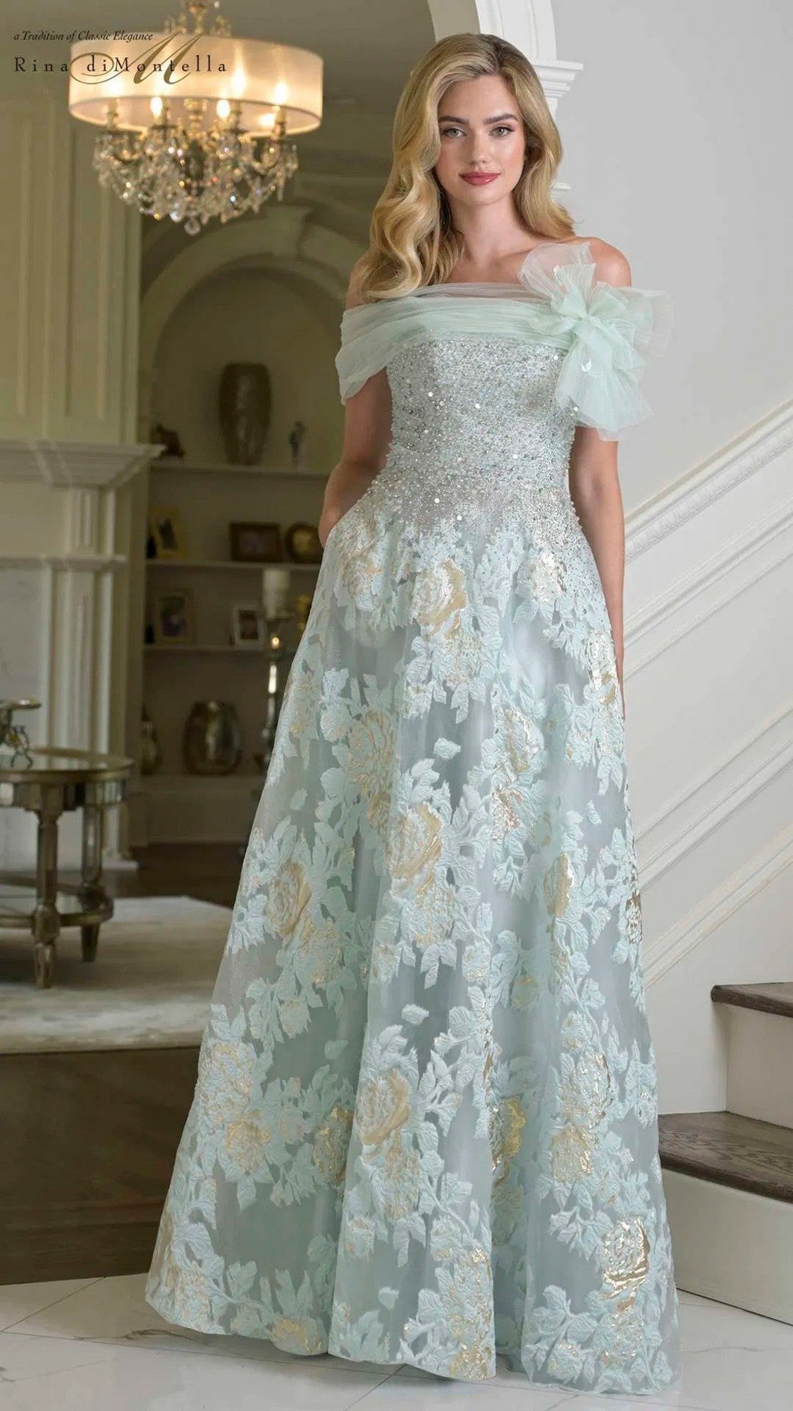A woman in the Rina Di Montella RD3406 long formal dress in seaglass, featuring floral embroidery and an elegant A-line skirt, stands indoors with a sparkling chandelier in the background.